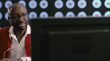 Kel Afro-Amerikalı iş adamı, kırmızı takım elbise, beyaz gömlek, şık gözlükler ve beyaz telefon kulaklığı giyiyor, bilgisayar başında müzik dinliyor, karanlık odada klasik arka planda oturuyor..