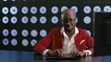 Olgun görünümlü, kırmızı takım elbiseli, beyaz gömlekli, şık gözlükler ve telefon kulaklıklı, gülücükler, klavyedeki tipler, müzik dinliyor, şarkı söylüyor, mavi duvarlı karanlık bir odada oturuyor..