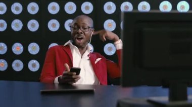 Kırmızı takım elbiseli, beyaz gömlekli, şık gözlüklü, telefon kulaklıklı, gülümseyen, masaüstünde oturan, müzik dinleyen, şarkı söyleyen, mavi duvarları olan, enerjik, Afro-Amerikalı olgun bir adam..