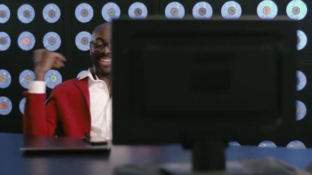 赤いスーツ 白いシャツ スタイリッシュなメガネと携帯電話のヘッドセット デスクトップに座って 音楽に耳を傾け 青い古典的な壁で暗いオフィスで踊る動きを持つ精力的なアフリカ系アメリカ人ビジネスマン — ストック動画