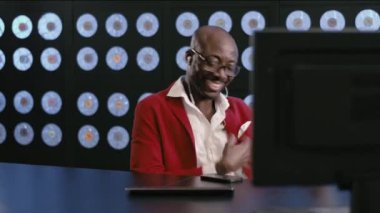 Sakallı, Afro-Amerikalı, kırmızı takım elbiseli, beyaz gömlekli, şık gözlükleri ve kulaklıklı, gülümseyen, masaüstünde oturan, müzik dinleyen, karanlık ofiste müzik aleti çalan,.