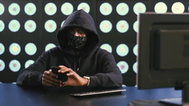 美国黑人男性计算机安全黑客 头戴黑脸面具 头戴黄帽 坐在黑暗的现代房间里 手握智能手机 看着它 — 图库视频影像