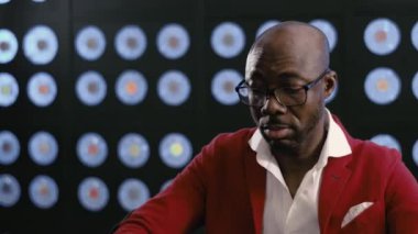 Kırmızı takım elbiseli, beyaz gömlekli ve şık gözlüklü profesyonel bir Afro-Amerikan işadamı bilgisayarda çalışıyor. Karanlık bir ofiste mavi klasik rengin bulanık arka planıyla oturuyor..