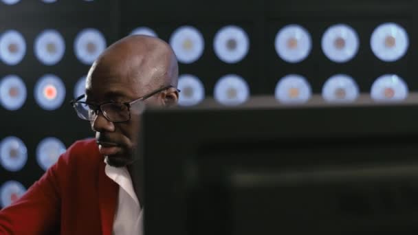 成熟した髭を生やしたアフリカ系アメリカ人ビジネスマン 赤いスーツを着て 白いシャツとスタイリッシュな眼鏡を着用してコンピュータ上で作業している 青い古典的な色を持っている背景がぼやけて暗い部屋に座っている — ストック動画