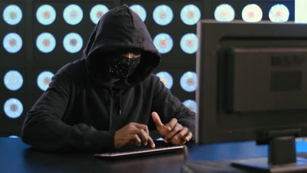看到年轻的非洲裔美国男性电脑黑客戴着黑色的面具和帽衫坐在黑暗的现代房间里 积极地在键盘上打字 凝视着橱窗 — 图库视频影像
