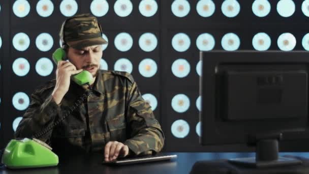 身穿迷彩服 头戴便帽的军人坐在办公桌前 手持电脑 用黑色无线键盘打字 用绿色老式电话交谈 房间漆黑 墙壁蓝色经典 — 图库视频影像
