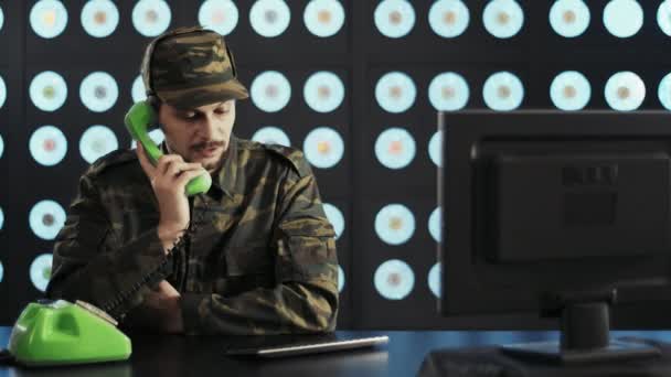 身穿军用迷彩服 头戴军帽的英俊大胡子男子与个人电脑坐在办公桌前 用绿色老式电话与蓝色经典墙纸在黑暗的现代房间里交谈 — 图库视频影像