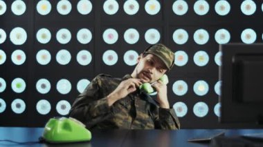 Askeri kamuflaj üniforması giymiş, sakallı ve şapkalı bir adam bilgisayar başında, kablosuz klavyenin yanında, modern duvarlı, yeşil, eski moda bir telefonla konuşuyor..