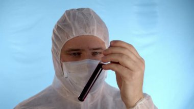 Beyaz koruyucu takım elbiseli, tıbbi maskeli, mavi arka planda lastik eldivenli beyaz bir adam kırmızı sıvı ve 2019 nCoV yazısıyla el sıkışarak test tüpünü tutuyor.