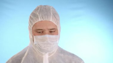 Beyaz koruyucu takım elbiseli, tıbbi maskeli, mavi arka planda lastik eldivenli beyaz bir adam kırmızı sıvı ve Coronovirus yazısıyla el tüpünü tutuyor ve kameraya test tüpünü gösteriyor.
