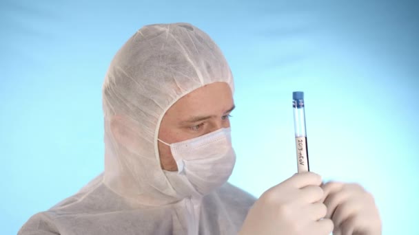 白い保護スーツ 医療用マスク 青の背景にゴム手袋の白人男性は 赤い液体と碑文コロノウイルスと手のテストチューブを保持し その後 カメラへのテストチューブを示しています — ストック動画