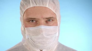 Beyaz koruyucu takım elbiseli beyaz adam, tıbbi maske, mavi arka planda lastik eldiven kırmızı sıvı ve 2019-nCoV yazısıyla el tüpü tutuyor, sonra da kameraya test tüpünü gösteriyor.