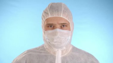 Beyaz koruyucu takım elbiseli beyaz bir adam, tıbbi maske, mavi arka planda lastik eldivenler kırmızı sıvı ve yazıt koronovirüs içeren test tüpü tutuyor, sonra da test tüpünü yere düşürüp kameraya bakıyor.