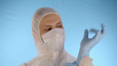 Beyaz koruyucu takım elbiseli ve mavi arka planda tıbbi maskeli beyaz bir adam ellerinde lastik eldivenleri düzeltiyor..