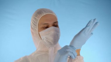 Beyaz koruyucu takım elbiseli ve mavi arka planda tıbbi maskeli beyaz bir adam ellerine lastik mavi eldiven takıyor.