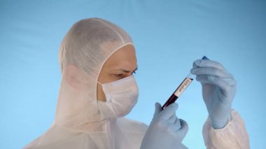 Beyaz koruyucu takım elbiseli, tıbbi maskeli, mavi arka planda lastik eldivenli beyaz bir adam kırmızı sıvı ve Coronovirus yazısıyla el tüpünü tutuyor ve kameraya test tüpünü gösteriyor.