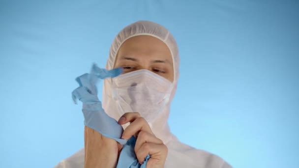 身穿白色防护服 戴着蓝色背景医疗面具的白人男子将橡胶蓝色手套戴在手上 — 图库视频影像
