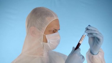 Beyaz koruyucu giysili adam, tıbbi maske, lastik eldiven kırmızı sıvı ve yazıt koronovirüs ile test tüpü tutuyor, terini peçeteyle siliyor, kameraya test tüpü gösteriyor.