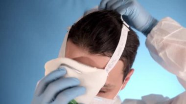 Mavi arka planda beyaz koruyucu takım elbiseli beyaz sakallı adam yüzüne solunum cihazı, gözlük ve iki tıbbi maske takıyor..