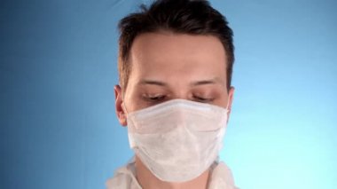 Mavi arka planda beyaz koruyucu takım elbiseli beyaz sakallı adam yüzüne solunum cihazı, yüzüne iki sağlık maskesi, fermuar ceketi, kapüşon takıp sağ elini gösteriyor.