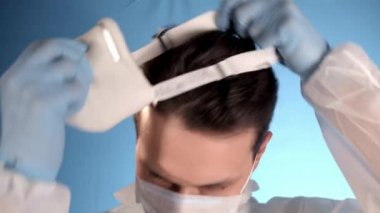 Mavi arka planda beyaz koruyucu takım elbiseli beyaz sakallı adam yüzüne solunum cihazı, yüzüne iki sağlık maskesi, fermuar ceketi, kapüşon takmış ve sol el gibi bir şey gösteriyor.