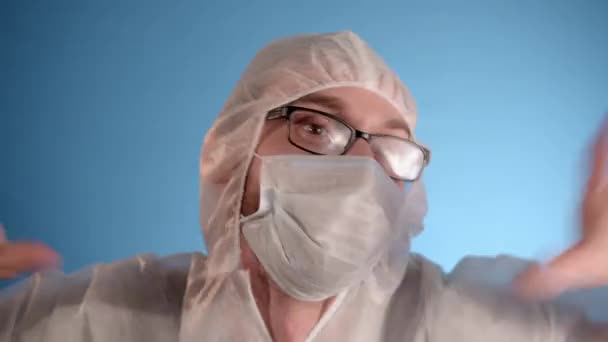 身穿白色防护服 戴着眼镜 戴着医疗面具的白人男子在蓝色背景的摄像机前跳舞 — 图库视频影像