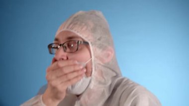 Beyaz önlüklü, koruyucu maskeli, mavi lastik eldivenli beyaz erkek laboratuvar teknisyeni mavi arka planda virüs kapmış, derin öksürükler ve dans.