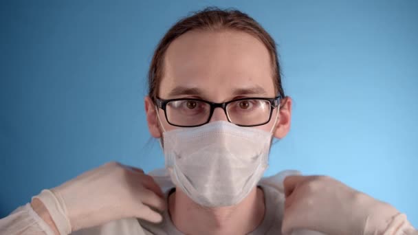 长发白胡子男子身穿蓝色背景的白色防护服 戴着眼镜 戴上医疗面罩 拉上夹克 戴上头罩 — 图库视频影像