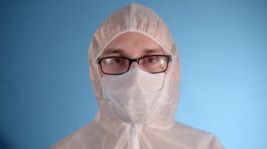 Mavi arka planda beyaz koruyucu tulumlu beyaz gözlüklü, uzun saçlı beyaz sakallı adam tıbbi maskeyi çıkartıyor, ceketinin fermuarını açıyor, derin derin iç çekiyor.