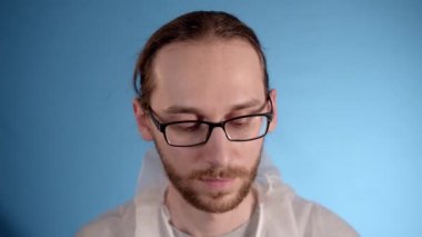 Uzun saçlı beyaz sakallı, gözlüklü bir adam mavi bir arka planda beyaz koruyucu bir tulum giyiyor. Panikle yüzüne bir tıbbi maske takıyor, ceketinin fermuarını çekiyor ve kapüşonunu takıyor..