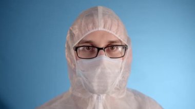 Beyaz koruyucu takım elbiseli, tıbbi maskeli, gözlüklü, mavi arka planda lastik eldivenli beyaz bir adam kırmızı sıvı ve Coronovirus yazısıyla el tüpünü tutuyor.