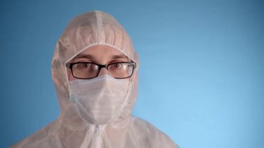 Beyaz koruyucu takım elbiseli, tıbbi maskeli, gözlüklü, mavi arka planda lastik eldivenli beyaz bir adam kırmızı sıvı ve yazıt virüsü ile el tüpünde tutuyor. Sonra açmak ve şaşırtmak.
