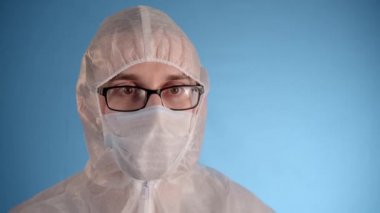 Beyaz koruyucu takım elbiseli, tıbbi maskeli, gözlüklü, mavi arka planda lastik eldivenli beyaz bir adam kırmızı sıvı ve yazıt virüsü ile el tüpünde tutuyor. Sonra aç, şaşırt ve kokla.