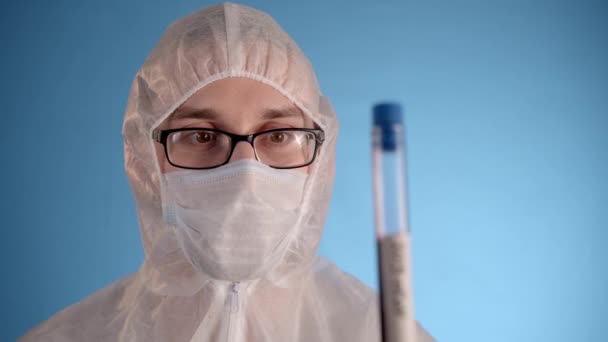 青い背景に防護服 医療用マスク ゴム手袋の白人男性は 赤い液体と碑文コロノウイルスと手のテスト管に保持しています 驚くべきことに匂いを嗅ぐと — ストック動画
