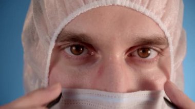 Beyaz önlüklü beyaz erkek laboratuvar teknisyeni, mavi arka planda koruyucu maske. Gözlükleri kapat.