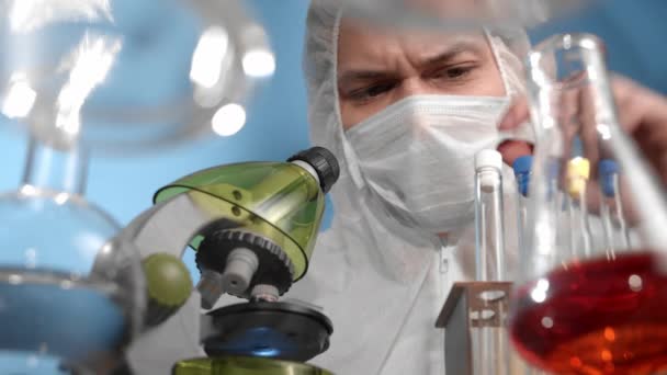 医療用マスクおよび保護全体の科学者は顕微鏡を通して見る ガラスの透明テーブルには 多色のソリューションを備えた容器とフラスコがあります 男はビールに赤い液体を注入している — ストック動画
