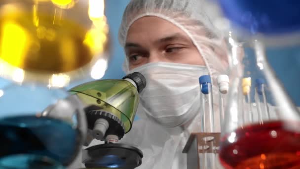 医療用マスクと保護ジャンプスーツの科学者が顕微鏡を調べる ガラスの透明テーブルには 多色のソリューションを備えた容器とフラスコがあります — ストック動画