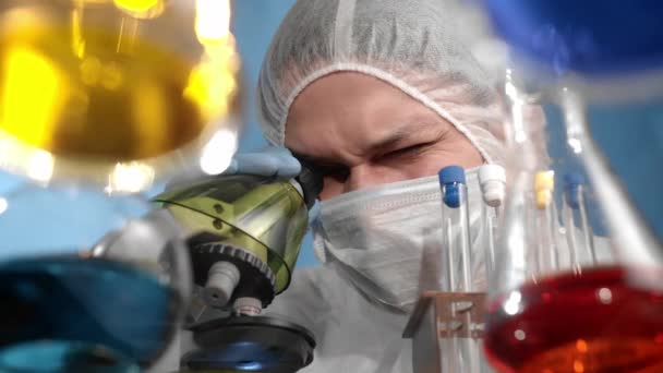 医療用マスクと保護ジャンプスーツの科学者が顕微鏡を調べる ガラスの透明テーブルには 多色のソリューションを備えた容器とフラスコがあります — ストック動画