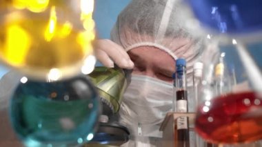Beyaz sakallı adam mikroskop tezgahına pipet yapıyor ve başarısız kimyasal deneyimi yüzünden üzgün. Laboratuvarı terk ediyor. Cam şeffaf masa üzerinde çok renkli çözeltileri olan mataralar var.