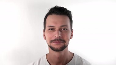 Beyaz tişörtlü, beyaz arka planda, stüdyoda sakallı, beyaz saçlı esmer bir adam gülümsüyor, kameraya doğru boğazdan konuşuyor ve gülüyor.