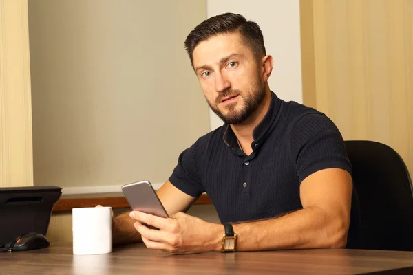 Hombre de negocios con taza y teléfono celular en la mano sentado a la mesa — Foto de Stock