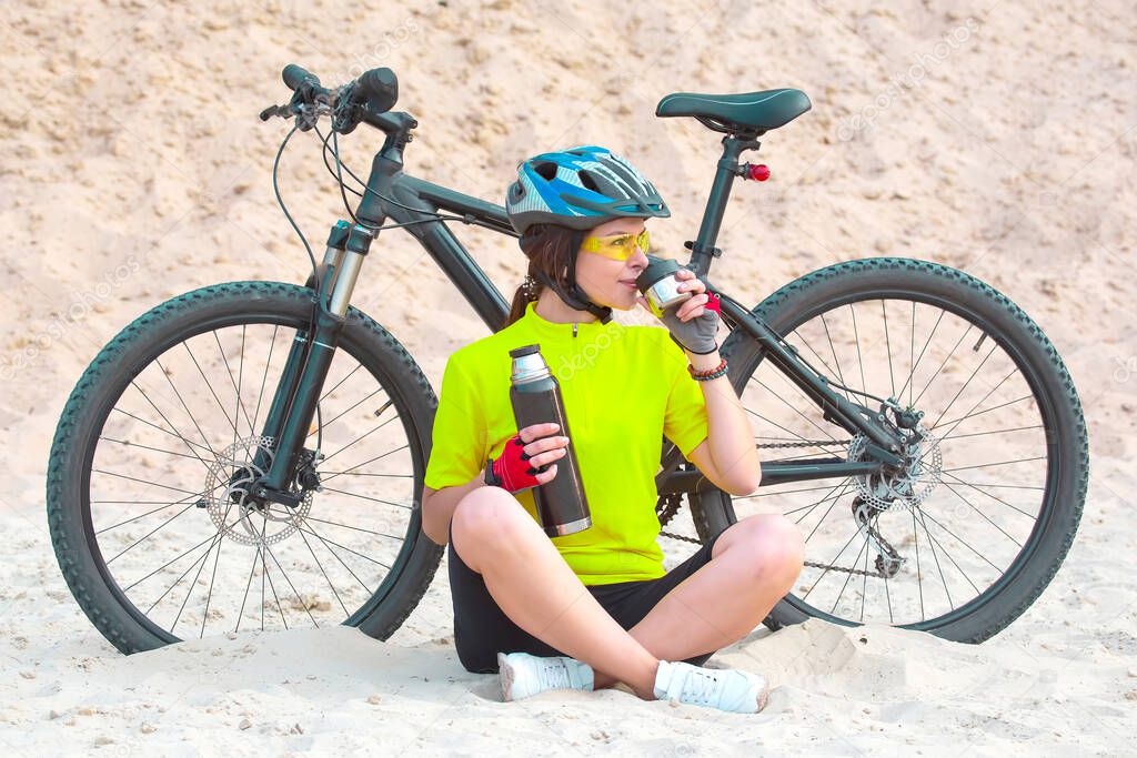 Beautiful girl cyclist pours tea from a thermos on the background of a Bicycle in the sand. Sports and recreation. Hobbies and health. Nature and man