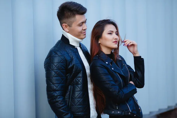 young asian guy and girl are standing in front of a light background. The girl leaned against the guy. Leather black jackets.