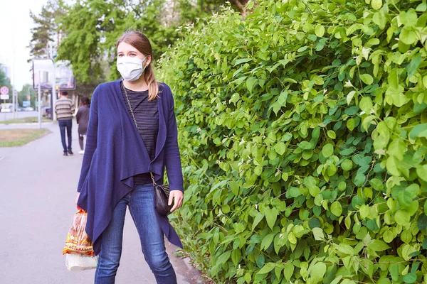 Mavi ceketli genç bir kız, tıbbi maske takan yeşil çalıların yanında duruyor.