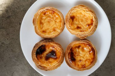 Dört Portekiz pastası, beyaz tabakta krema ve dokulu arka plan.