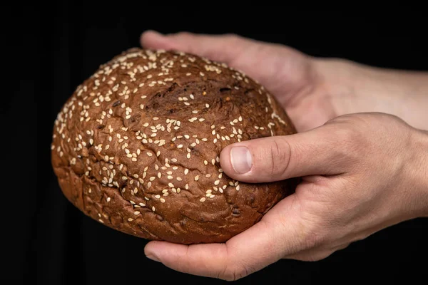 Man's hands hold tasty fresh loaf of dark bread with sesame seeds on black background — Stock Photo, Image