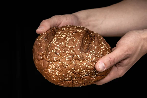 Man's hands hold tasty fresh loaf of dark bread with sesame seeds on black background — Stock Photo, Image