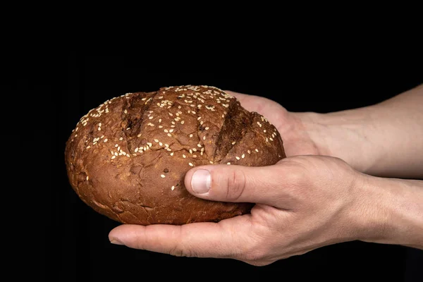 Man's hands hold tasty fresh loaf of dark bread with sesame seeds on black background — Stock Photo, Image
