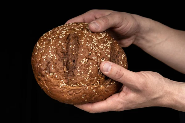 Man's hands hold tasty fresh loaf of dark bread with sesame seeds on black background — Stock Photo, Image