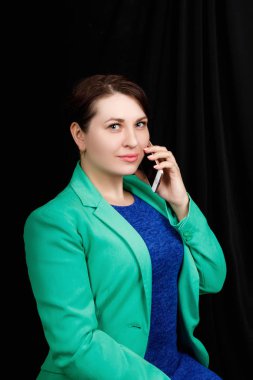 Studio  close up portrait of a girl of Slavic appearance worn a blue blouse and a blue-green jacket on a black background speaking by phone